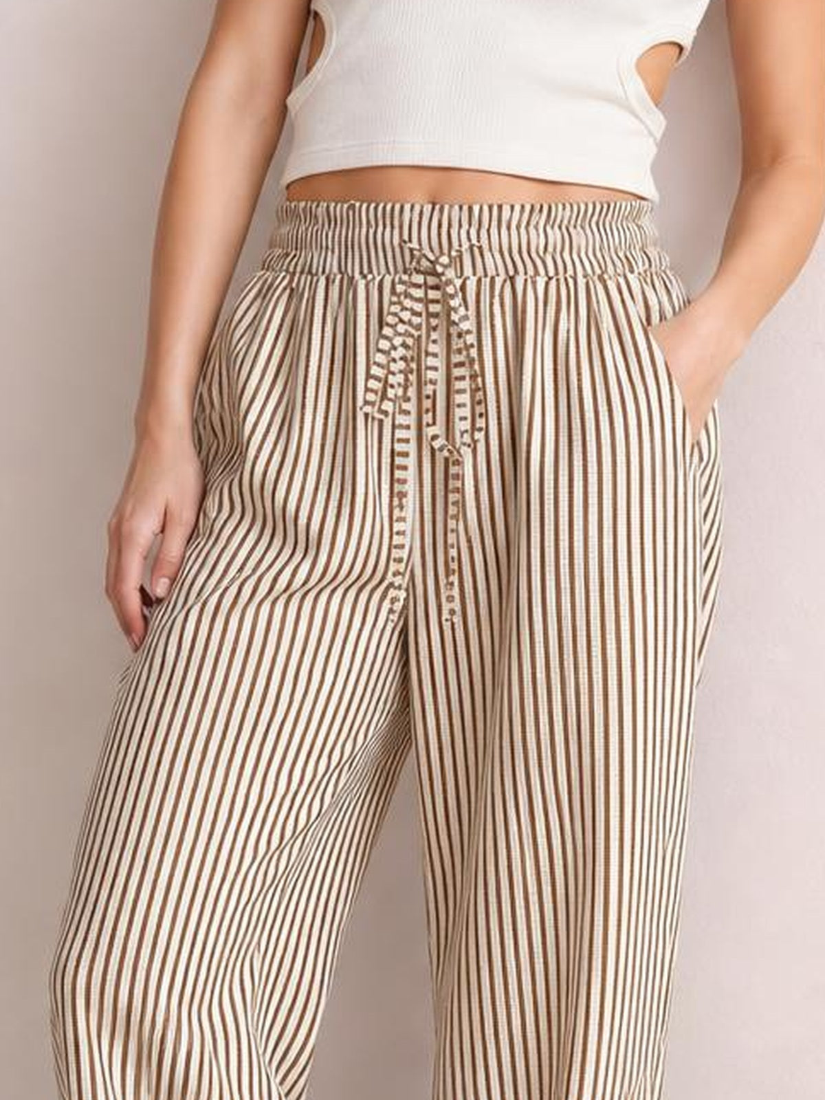 A cropped model wears cream and brown vertically striped pants with an elastic drawstring waist, relaxed wide legs, and hands tucked into the pockets.