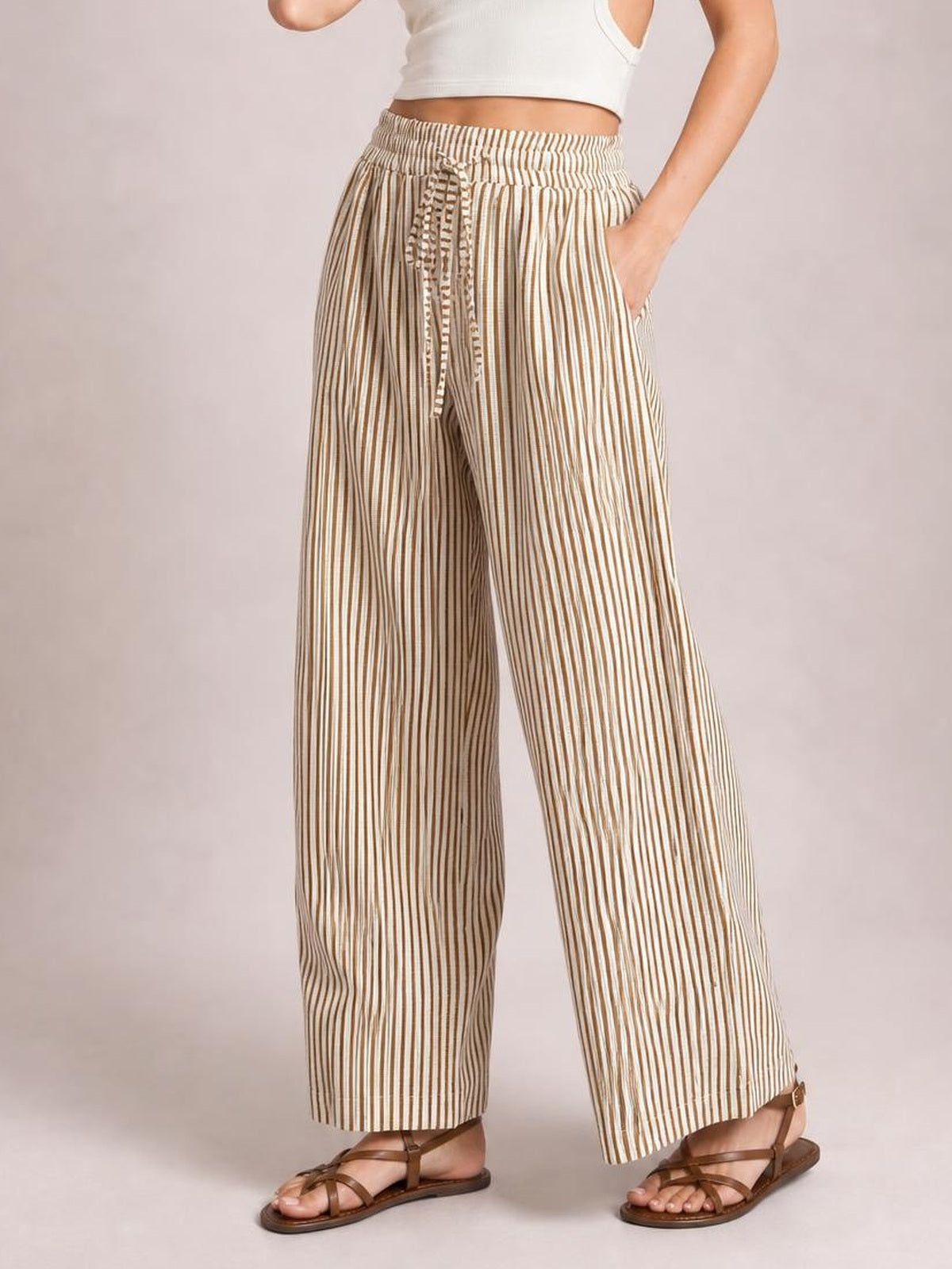 Model wearing tan and cream striped wide-leg pants with an elastic drawstring waist and side pockets, standing with one hand tucked in a pocket.
