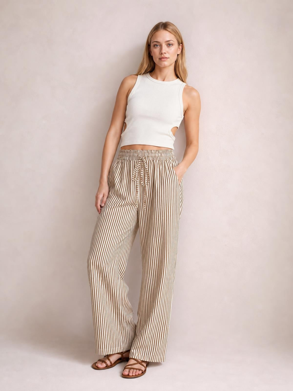 A model stands with one hand in a pocket, wearing beige-and-brown striped pants with an elastic drawstring waist and relaxed wide-leg silhouette.