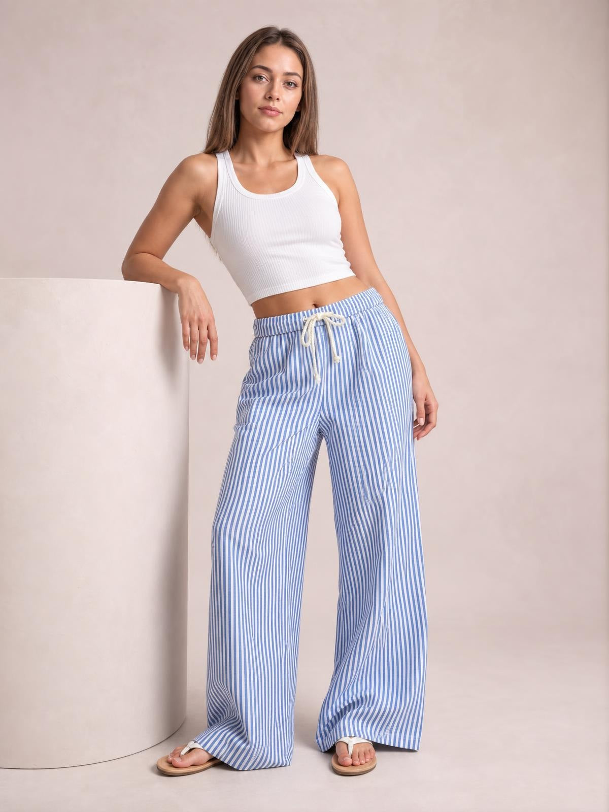 Model leans against a pedestal wearing blue-and-white striped wide-leg pants with a drawstring waist and relaxed, floor-skimming silhouette.