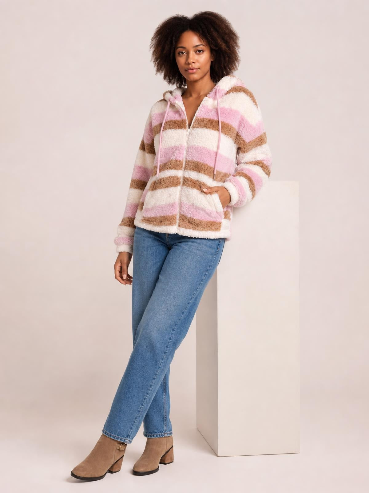A model poses in a plush pink, white, and brown striped zip-up hooded jacket with drawstrings, long sleeves, and a relaxed silhouette.
