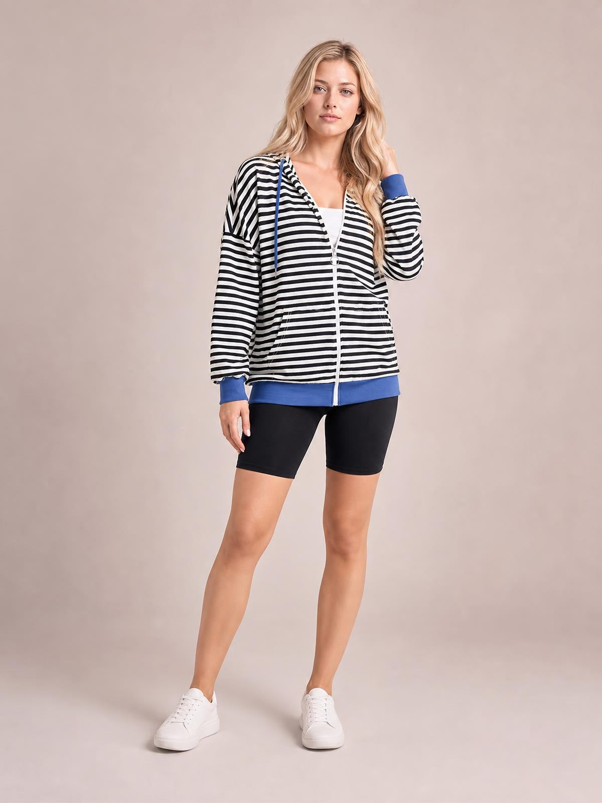 A woman stands in a relaxed pose wearing a black-and-white striped zip-up hoodie with blue drawstrings, cuffs, hem, and front pockets.