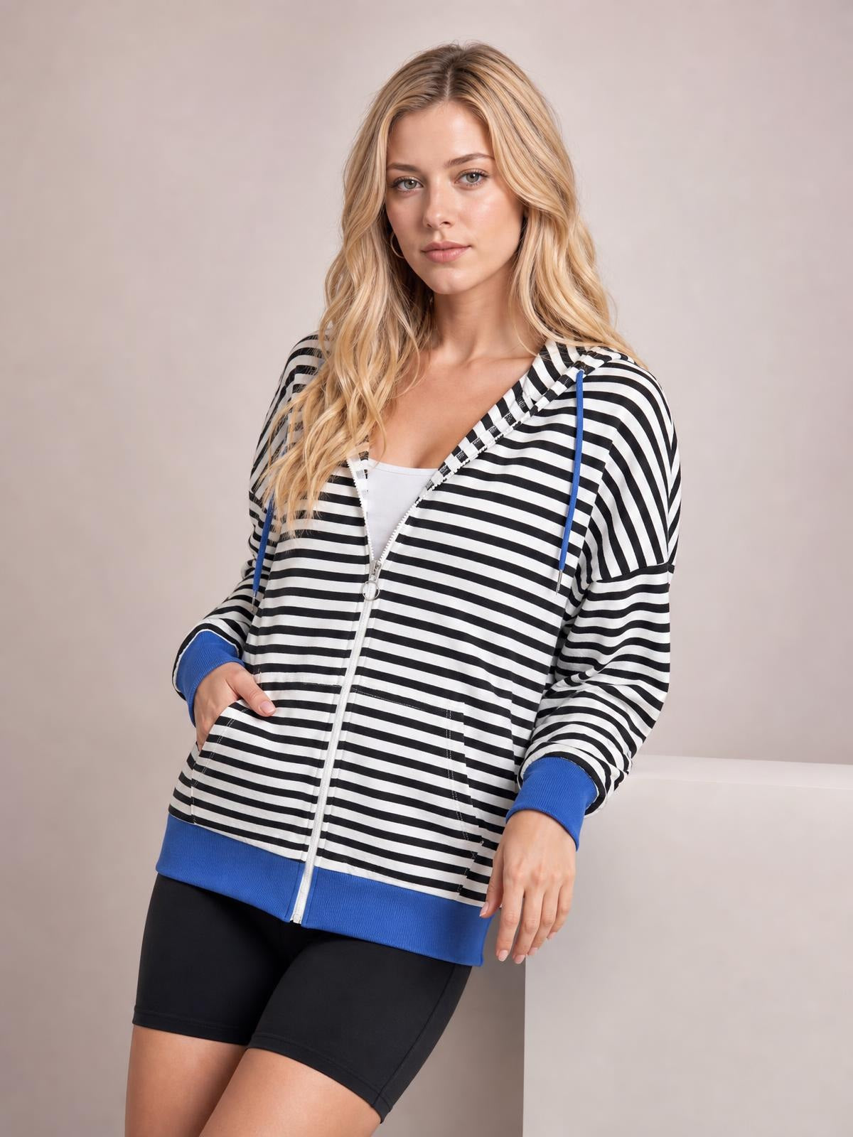 A model leans casually in a black-and-white striped zip-up hoodie with a drawstring hood, long sleeves, blue cuffs and hem, front pockets, and a relaxed silhouette.