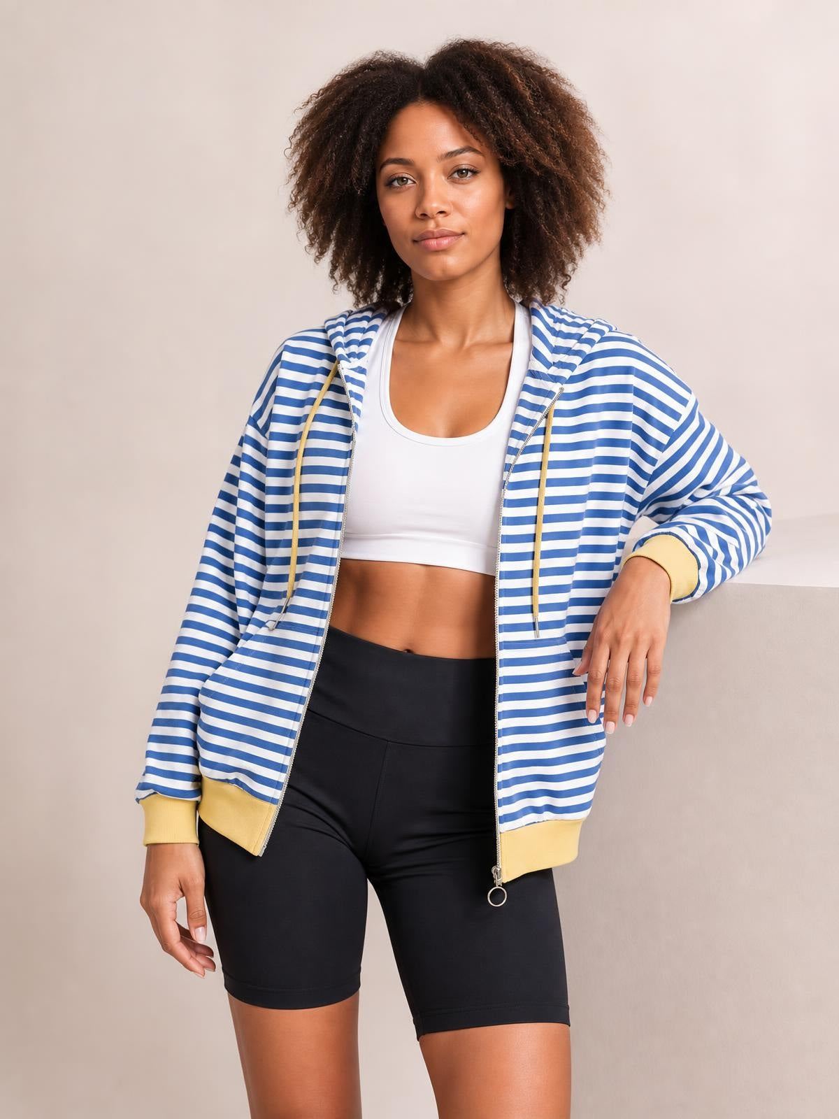 A model poses in a blue-and-white striped zip-up hoodie with drawstrings, long sleeves, a hood, yellow cuffs and hem, and a relaxed silhouette.
