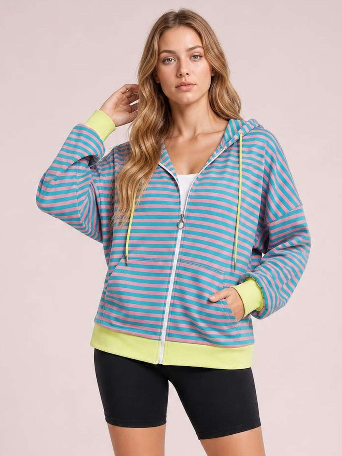 A model poses in a relaxed striped zip-up hoodie with turquoise and pink stripes, long sleeves, yellow drawstrings and cuffs, a hood, and front pockets.