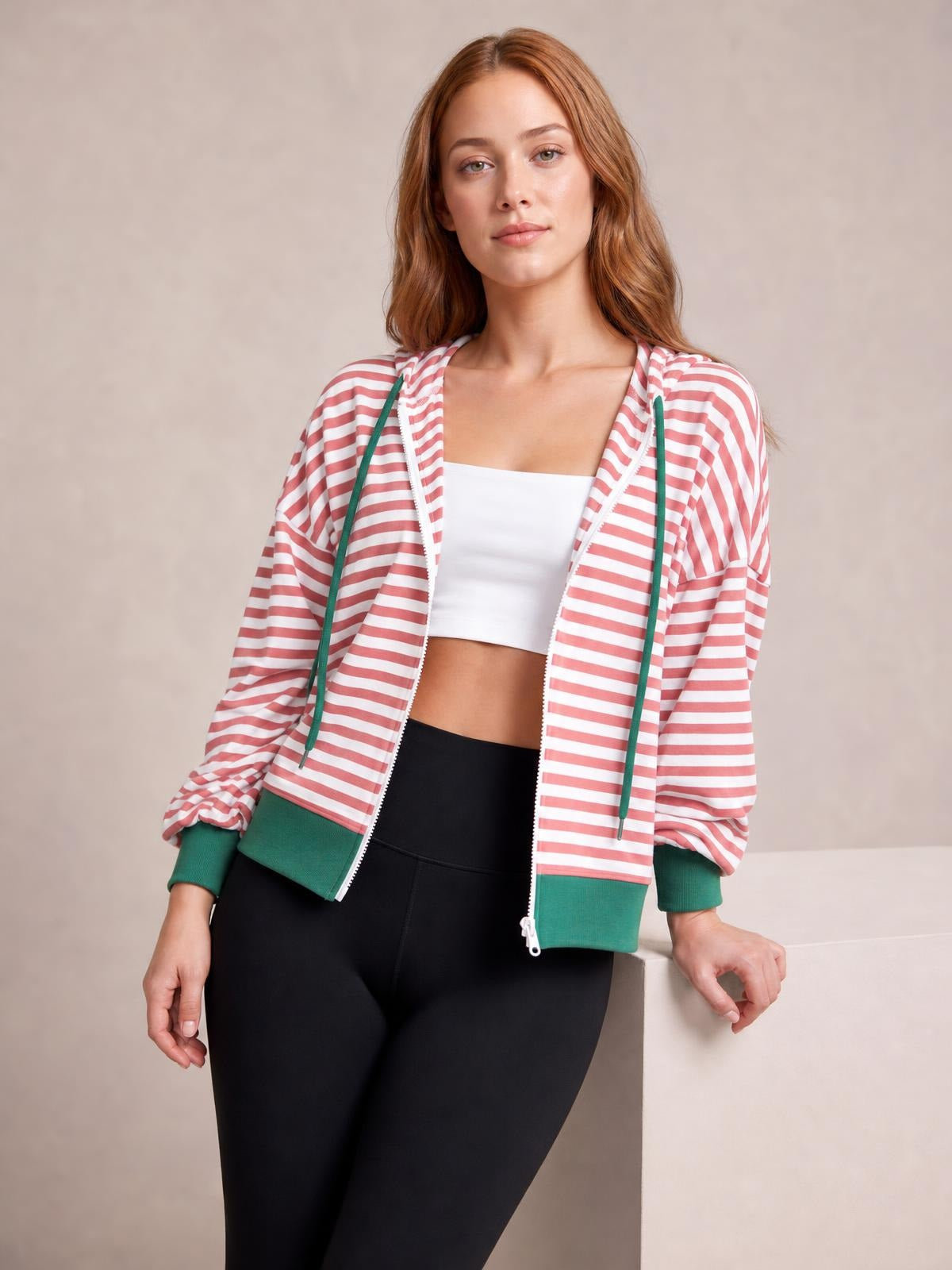 A model leans against a ledge wearing a pink-and-white striped zip-up hoodie with long sleeves, green drawstrings, ribbed cuffs, and a relaxed silhouette.