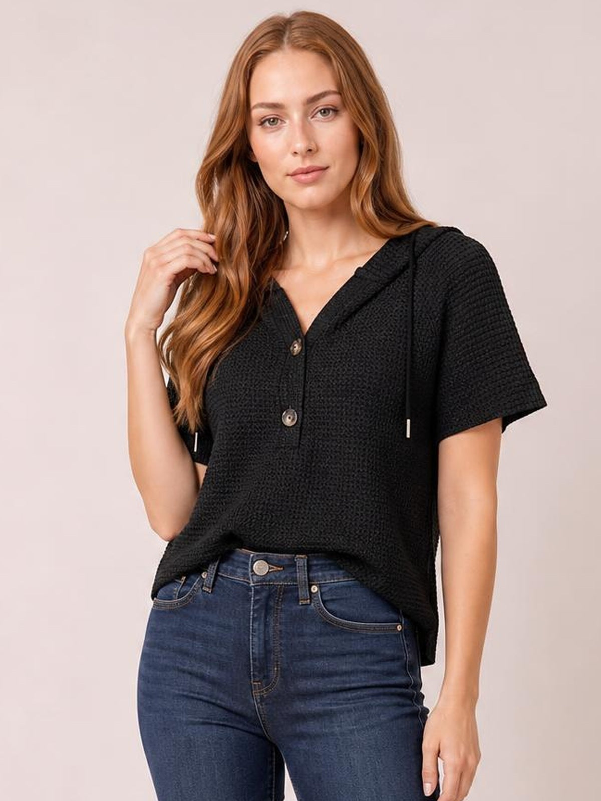 Model wears a black waffle-knit short-sleeve hooded top with drawstrings, buttoned front, and a relaxed silhouette while posing with one hand in her hair.
