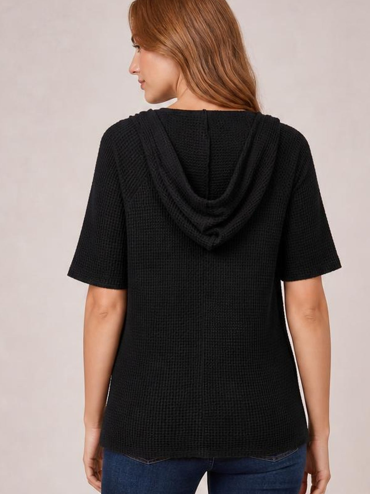 Back view of a model wearing a black waffle-knit short-sleeve hooded top with a draped hood and relaxed, slightly loose silhouette.