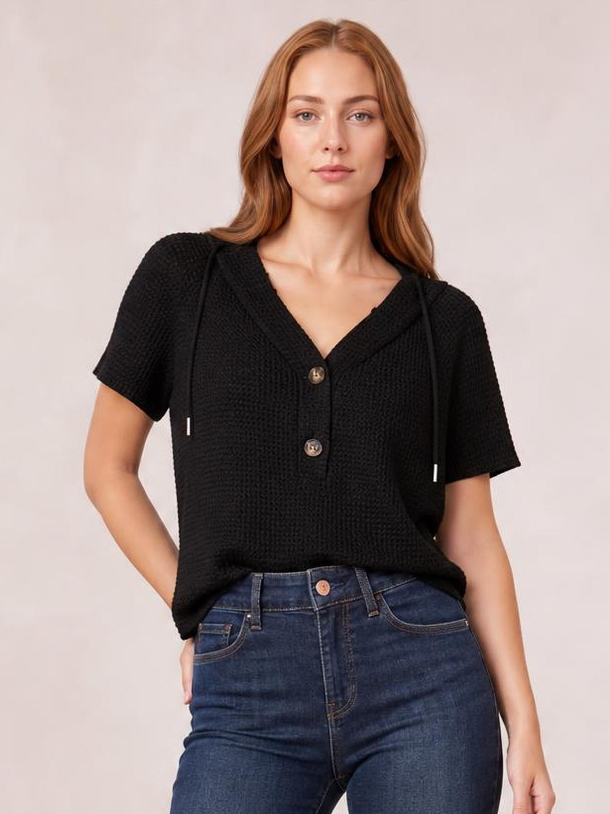 Model wears a black waffle-knit short-sleeve hooded top with drawstrings, button front, and relaxed silhouette, posing with one hand in her jeans pocket.