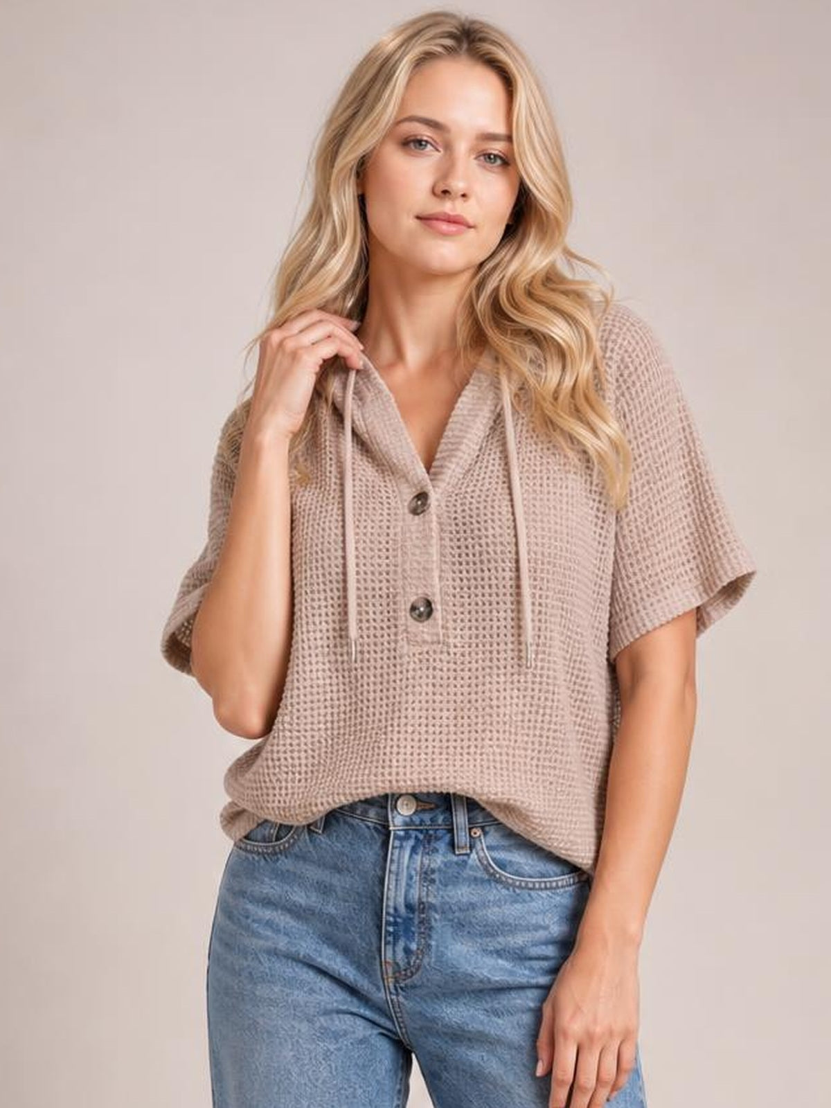 Model wearing a taupe waffle-knit short sleeve hooded top with drawstrings, buttoned front, and a relaxed silhouette, lifting the hood edge.