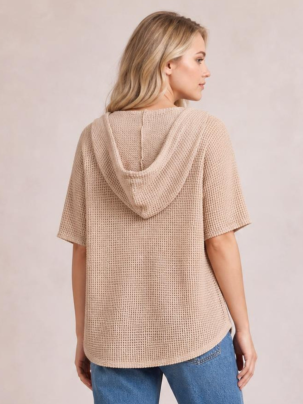 A woman models a beige waffle-knit short-sleeve hooded top with a relaxed straight silhouette and curved hem, shown from the back.