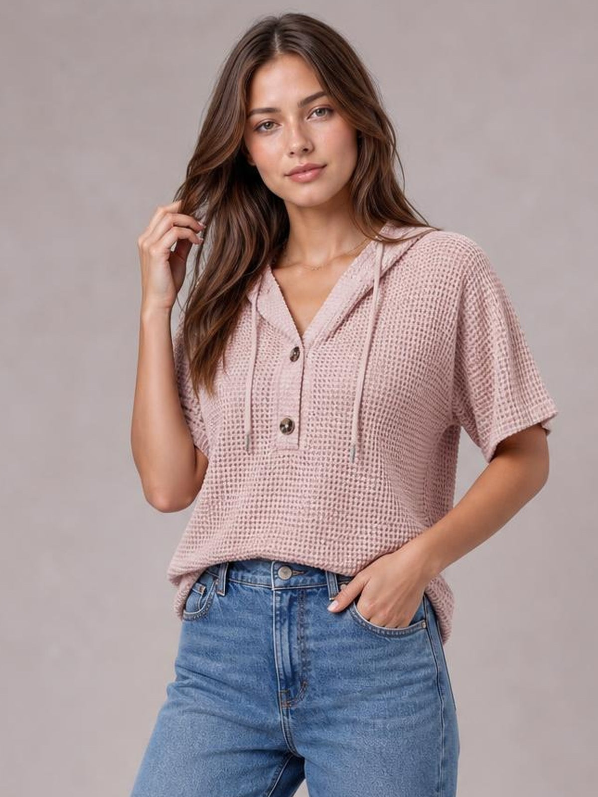 Model poses in a cozy blush pink waffle-knit short-sleeve hooded top with drawstrings, a buttoned placket, and a relaxed silhouette, paired with jeans.