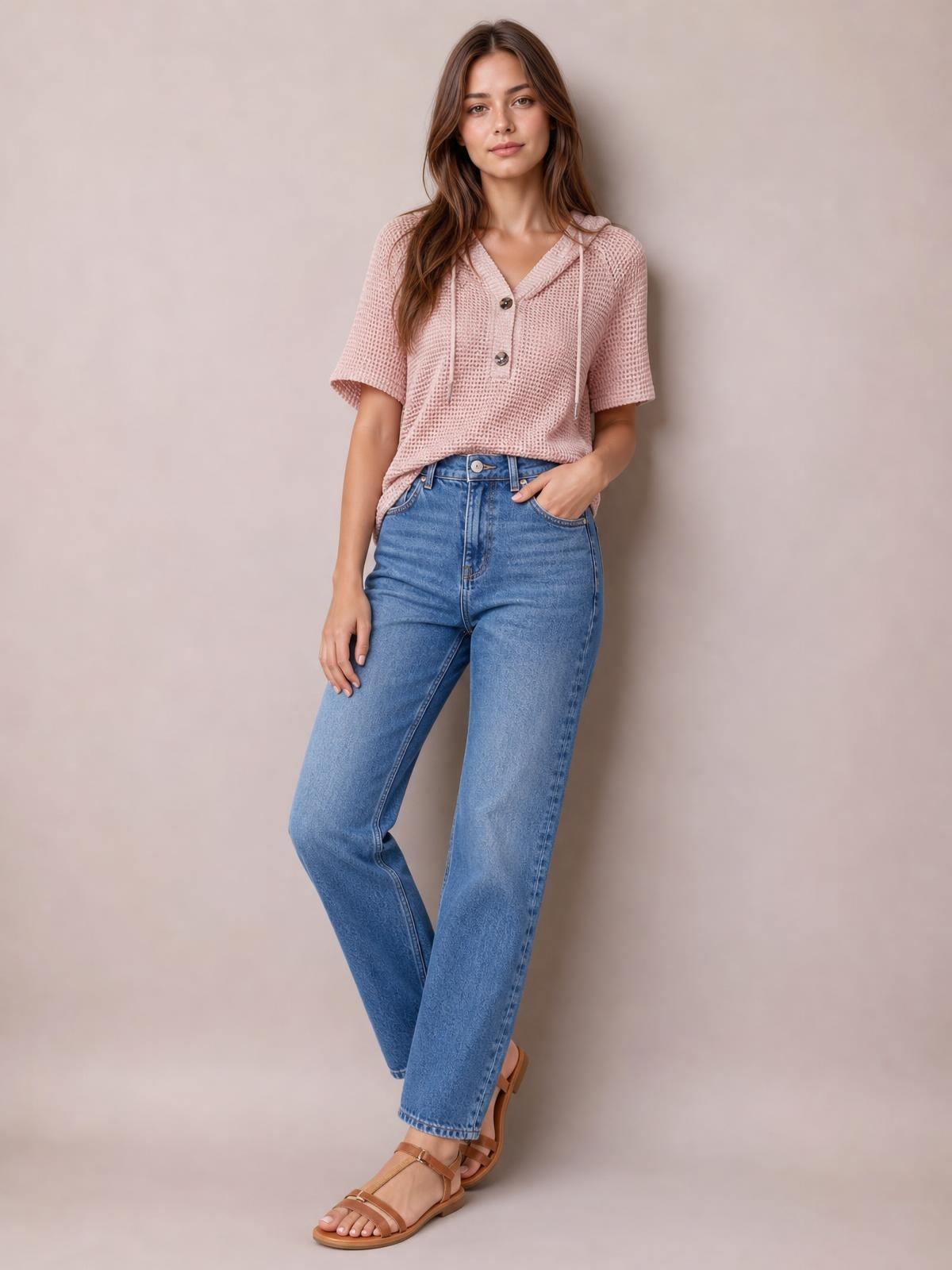 A model wears a dusty pink waffle-knit short-sleeve hooded top with drawstrings, a button placket, and a relaxed silhouette, posing with one hand in her jeans pocket.