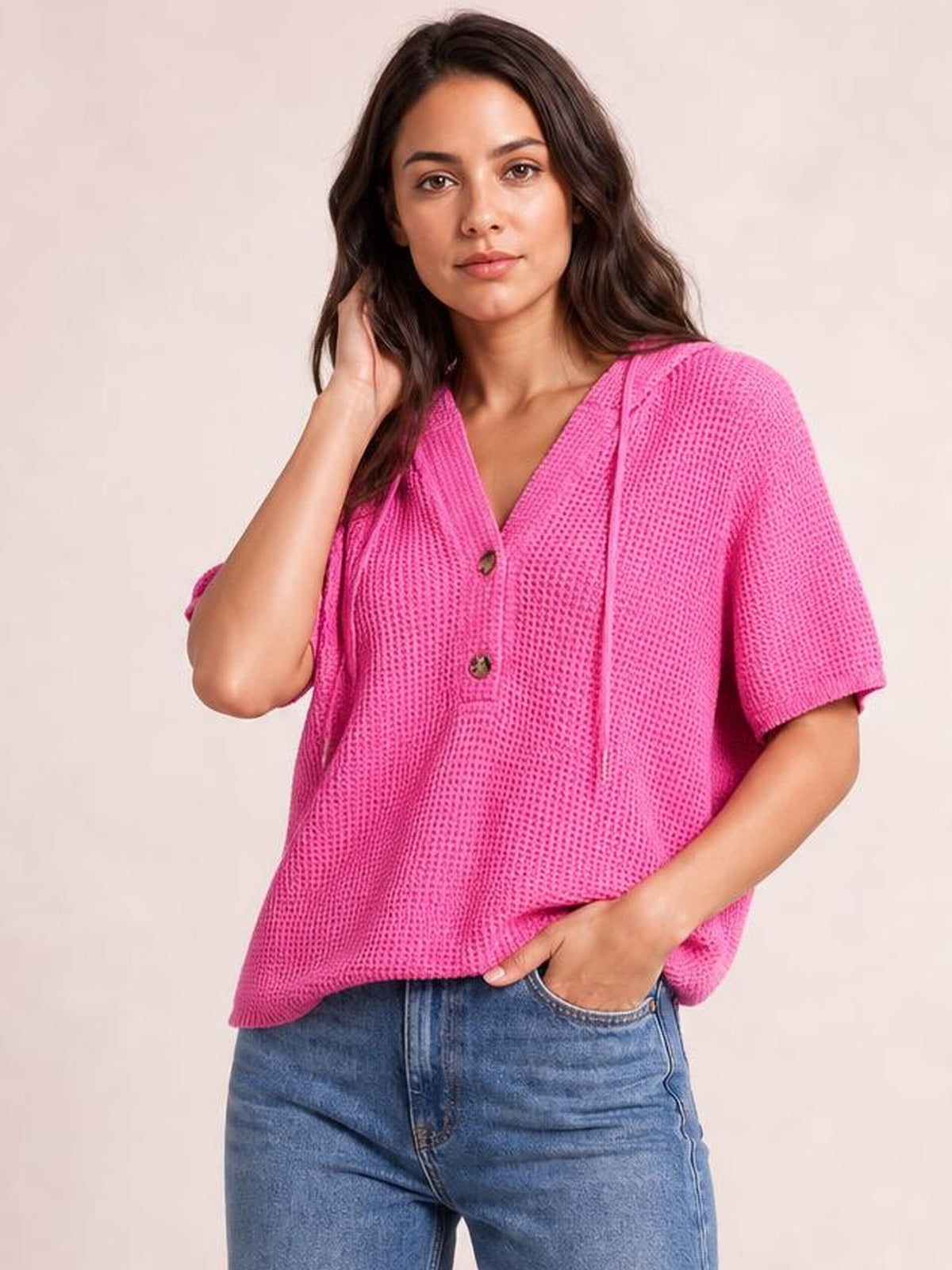 A model poses in a bright pink waffle-knit short-sleeve hooded top with drawstring ties, button front, and a relaxed silhouette.