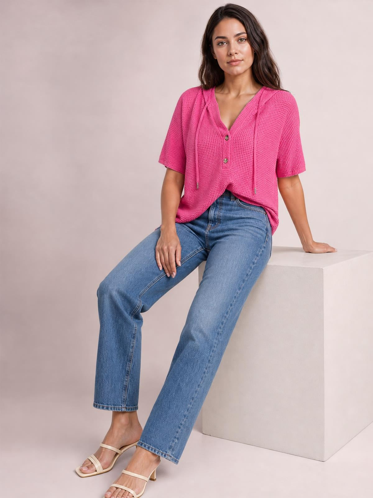 A seated model wears a bright pink waffle-knit hooded top with short sleeves, drawstrings, button front, and a relaxed silhouette.