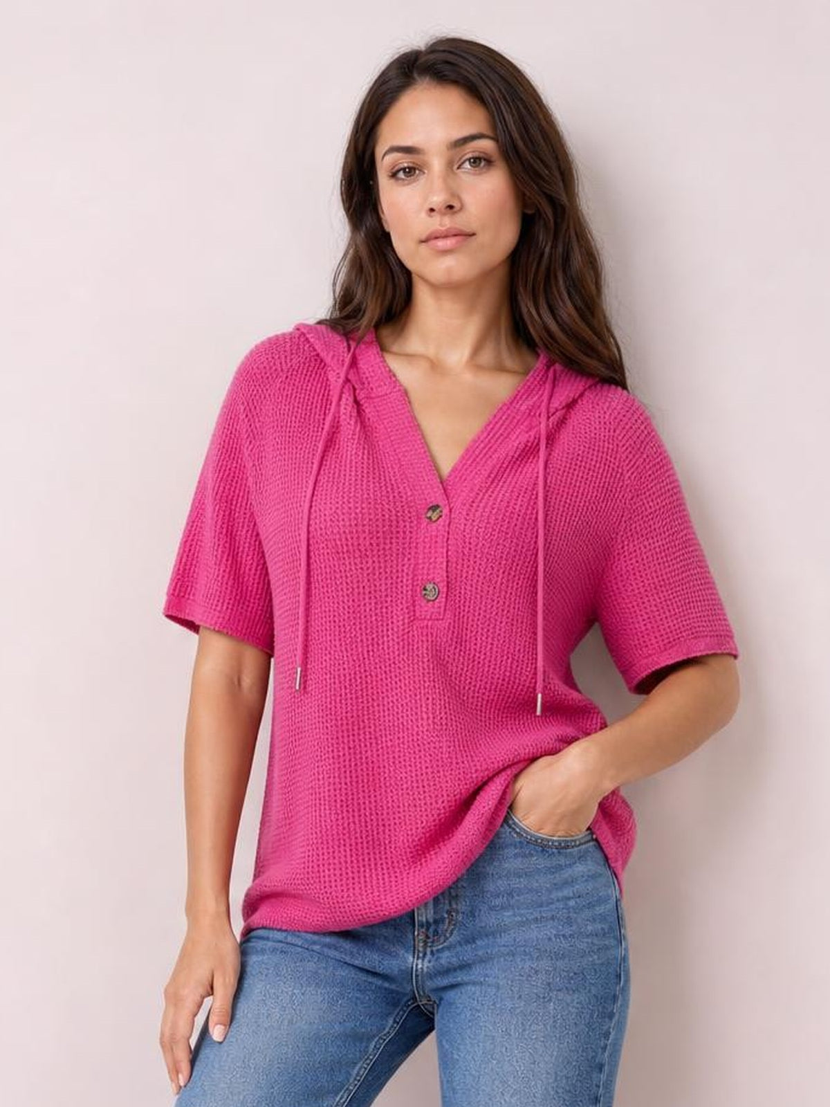 A model poses in a bright pink waffle-knit hooded top with short sleeves, drawstring hood, button placket, and a relaxed silhouette.