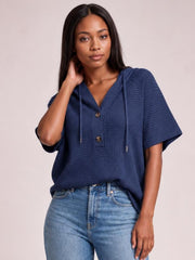 Model wearing a relaxed navy waffle-knit short sleeve hooded top with drawstrings and a button-front placket, posing with one hand near her hair.