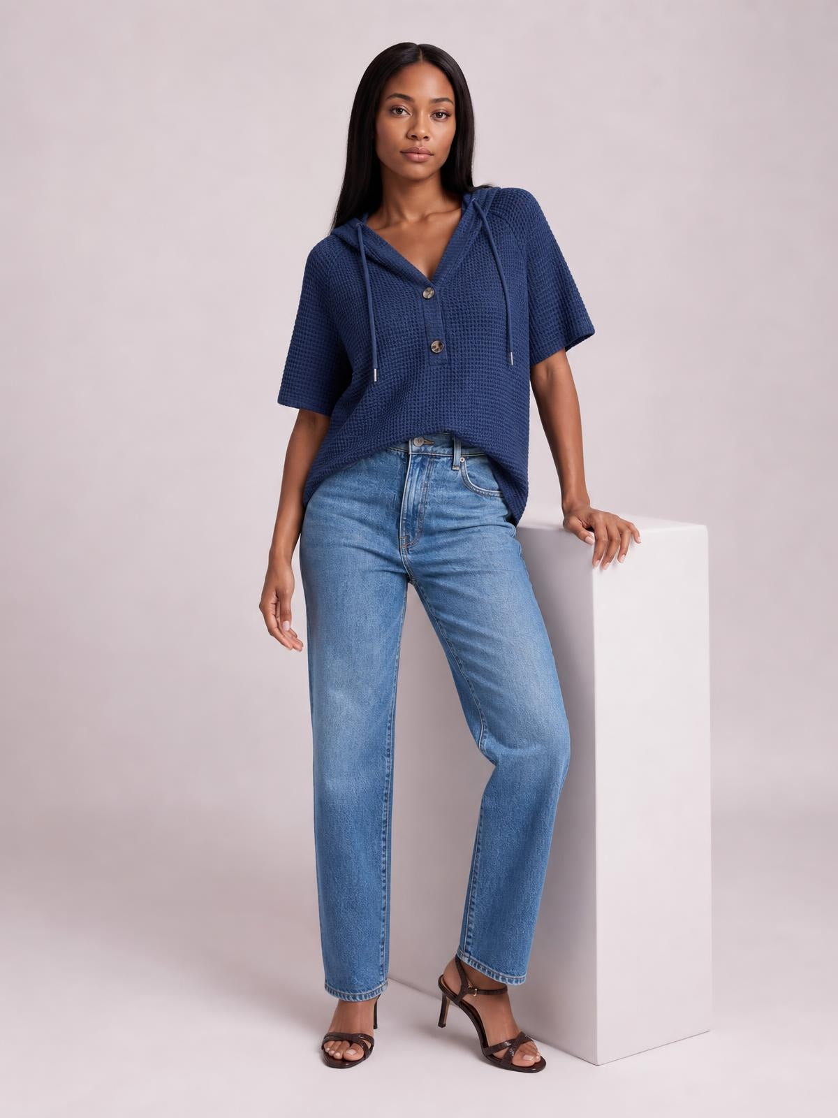 Model wears a navy waffle-knit short-sleeve hooded top with drawstrings and a two-button placket in a relaxed silhouette, leaning with one hand.