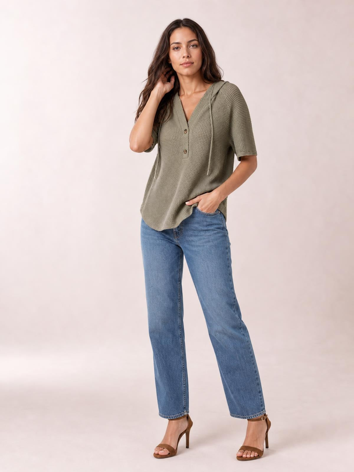 A model wears an olive waffle-knit short-sleeve hooded top with drawstrings, a button front, and a relaxed silhouette, posing with one hand in her pocket.