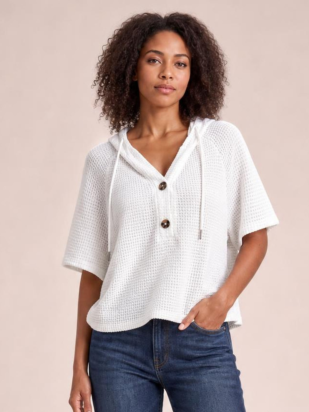 Model wears a white waffle-knit short-sleeve hooded top with drawstrings, buttoned front, and a relaxed silhouette while standing with one hand in her jeans pocket.