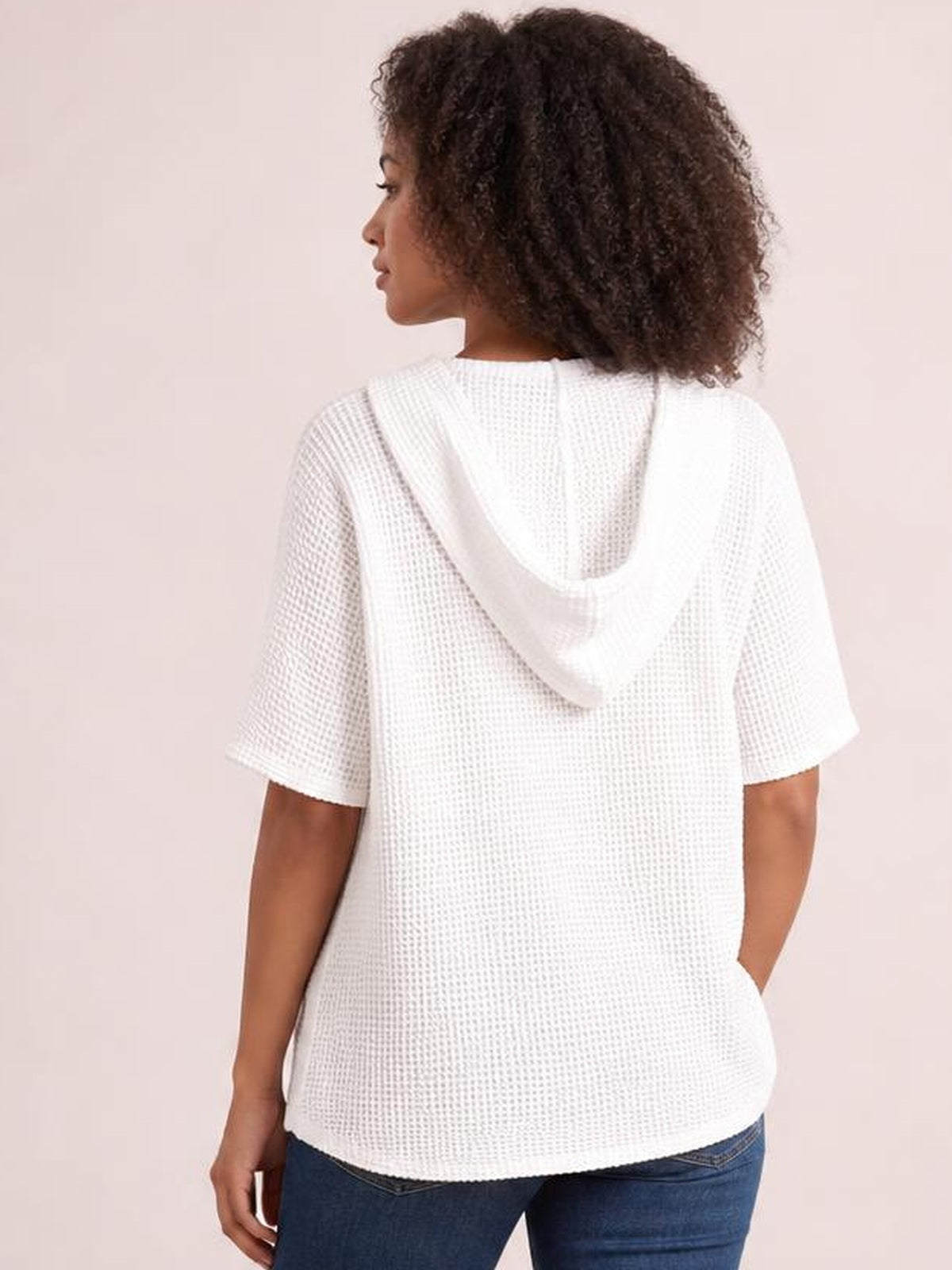 A model wears a white waffle-knit short-sleeve hooded top in a relaxed silhouette, shown from the back with one hand in a pocket.