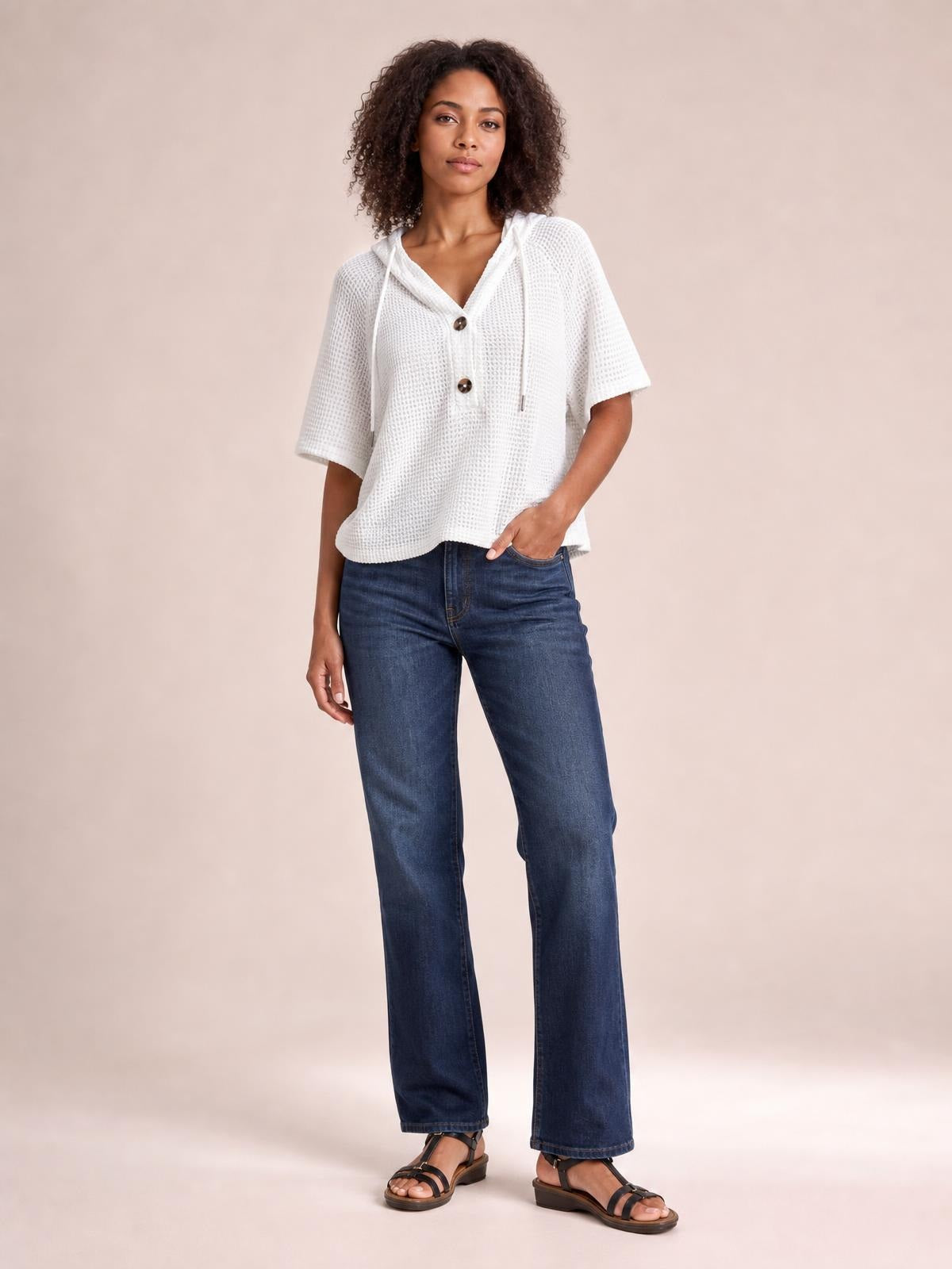 Model wears a white waffle-knit short sleeve hooded top with drawstrings, button front, and a relaxed silhouette, standing casually with one hand in her jeans pocket.