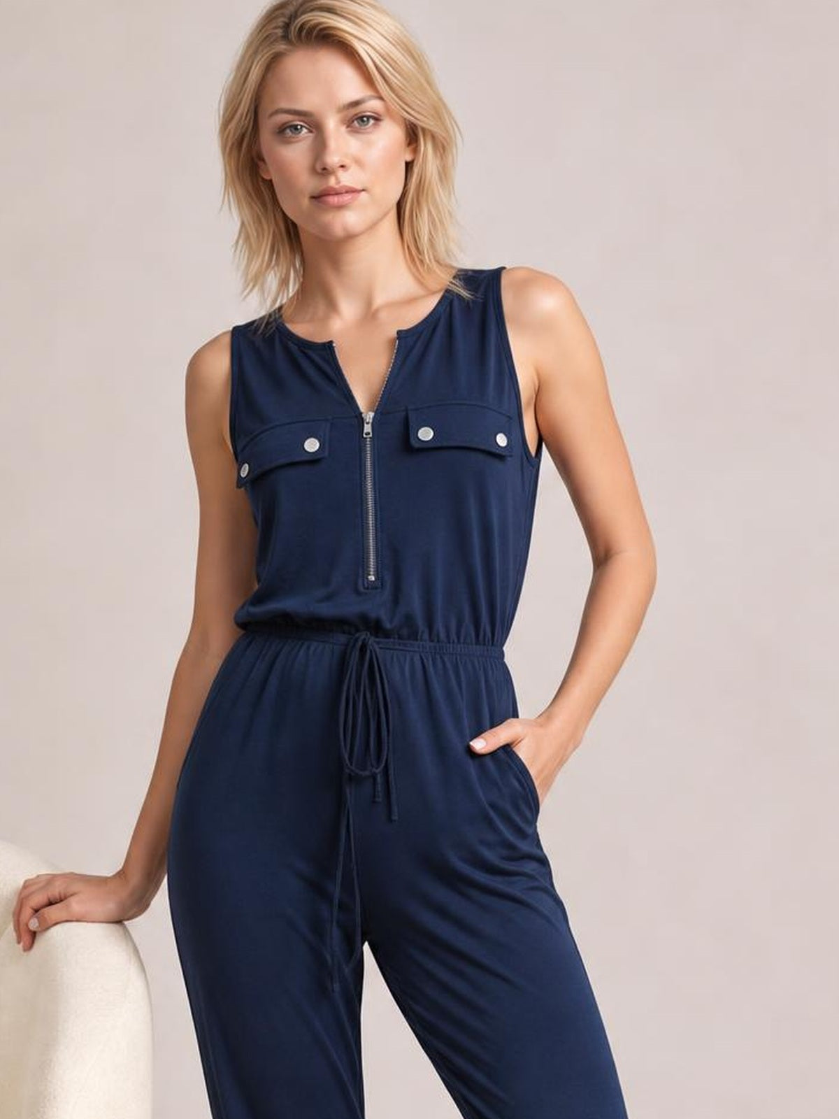 Model poses in a navy sleeveless jumpsuit with a front zipper, buttoned chest pockets, drawstring waist, and relaxed pocketed wide-leg silhouette.