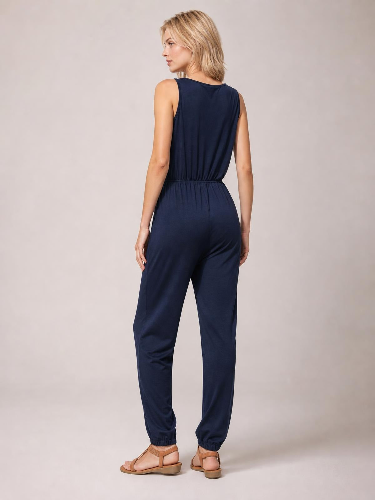 A model looks over her shoulder in a navy sleeveless jumpsuit with a gathered drawstring waist and relaxed tapered legs with elastic cuffs.