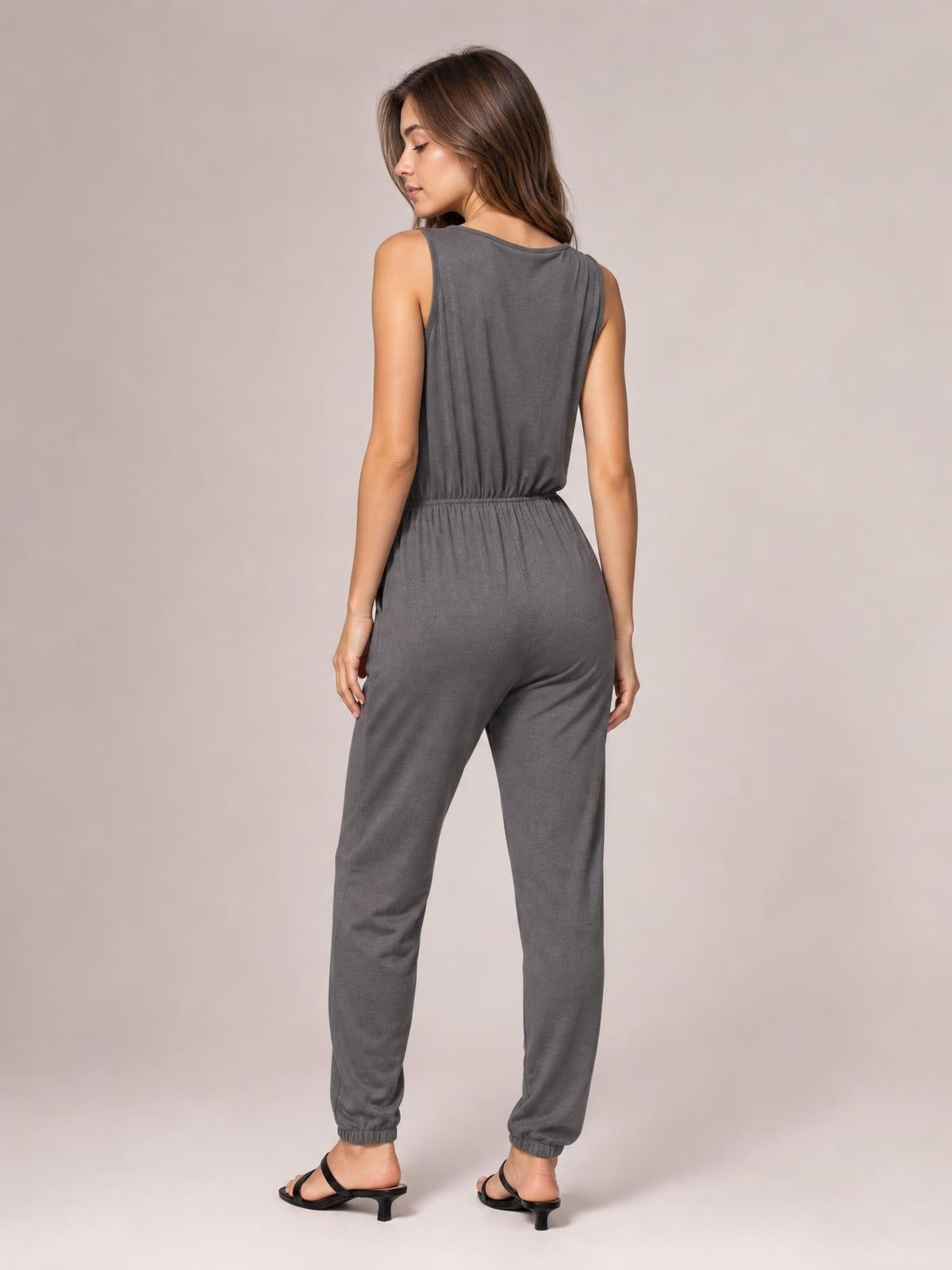 Woman looking over her shoulder in a charcoal gray sleeveless jumpsuit with a drawstring waist, relaxed tapered legs, and cuffed hems.