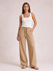 Model stands wearing tan drawstring wide leg pants with a relaxed full-length silhouette, elastic waistband, visible pockets, and one hand tucked into a pocket.