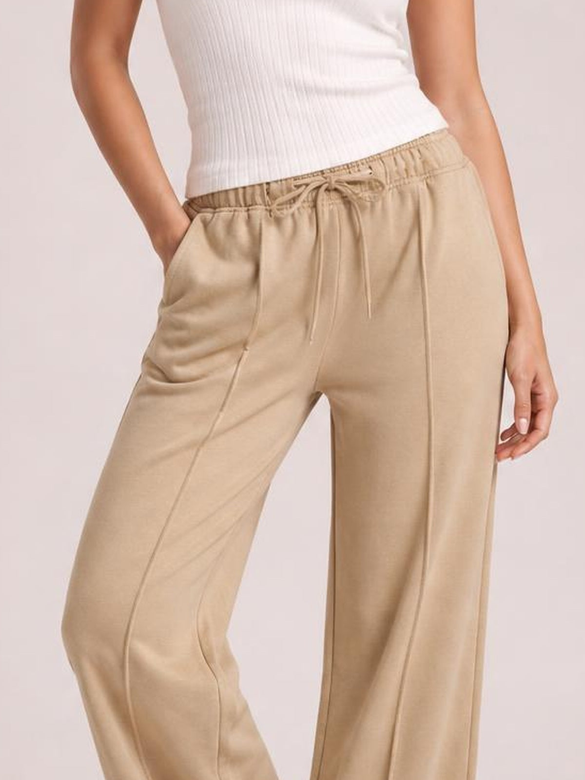 Model wearing beige drawstring wide leg pants with an elastic waist and side pockets, standing with one hand tucked into a pocket.