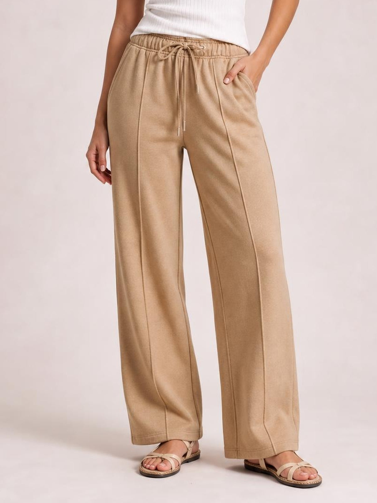 Model wears tan drawstring wide leg pants with an elastic waistband, visible side pockets, straight seams, and a relaxed silhouette while standing.