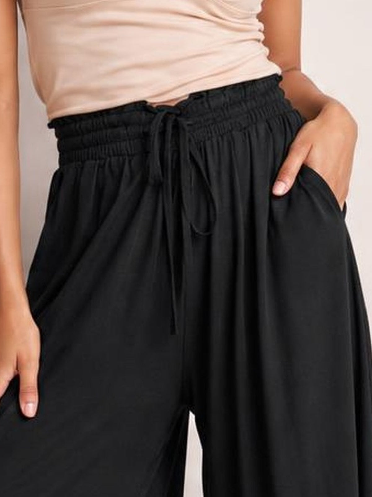 A cropped model wears black drawstring wide-leg pants with a gathered waistband, flowing silhouette, and roomy side pockets shown with hands tucked inside.