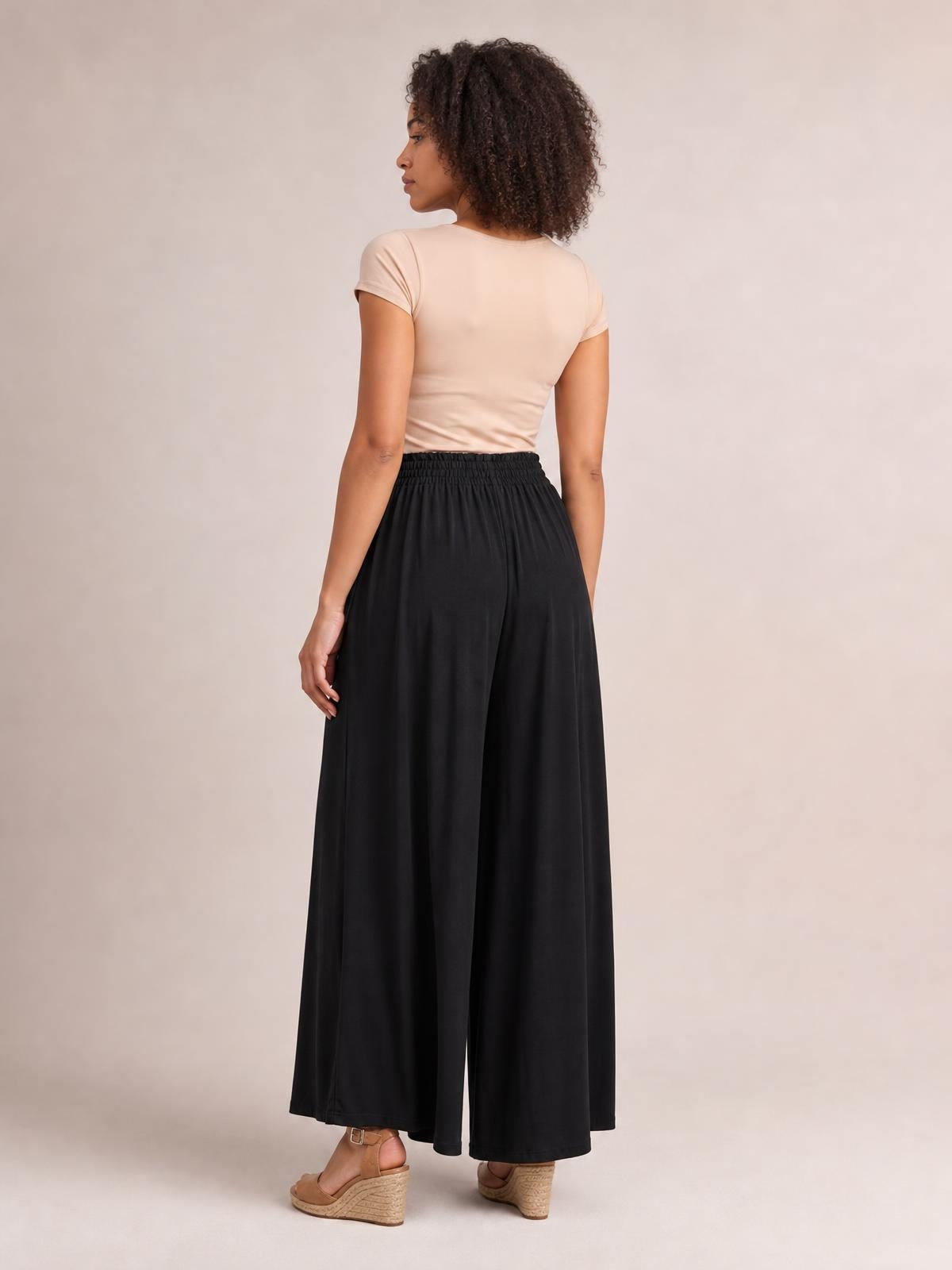 A model turns to show black drawstring wide-leg pants with a flowy, full-length silhouette, relaxed fit, and gathered elastic waistband.