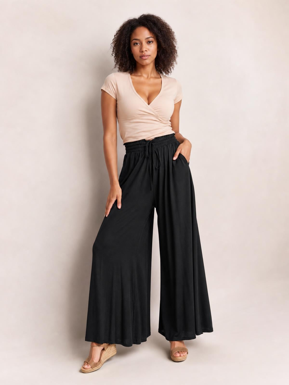 Model stands in black drawstring wide leg pants with side pockets, a flowy full-length silhouette, and one hand resting in a pocket.