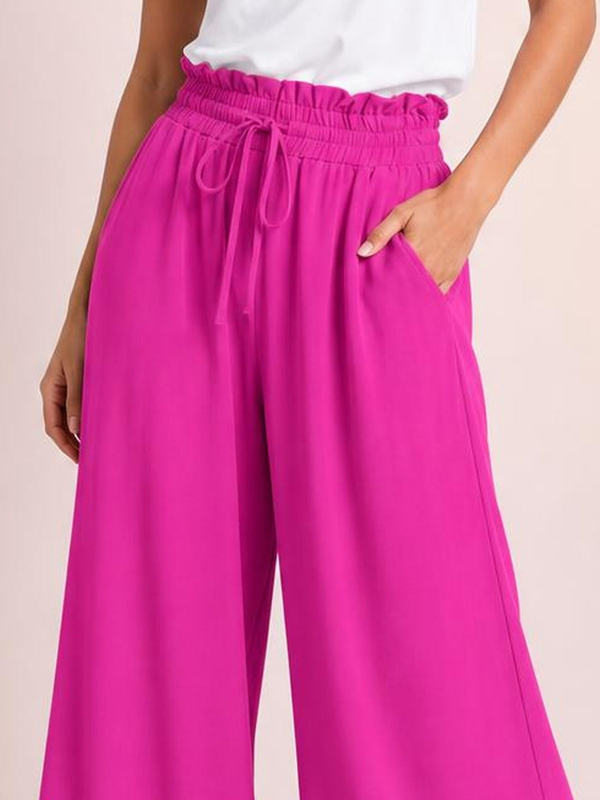 Model wears bright pink drawstring wide-leg pants with a flowy silhouette, elasticized waist, visible tie, and side pockets, one hand tucked inside.