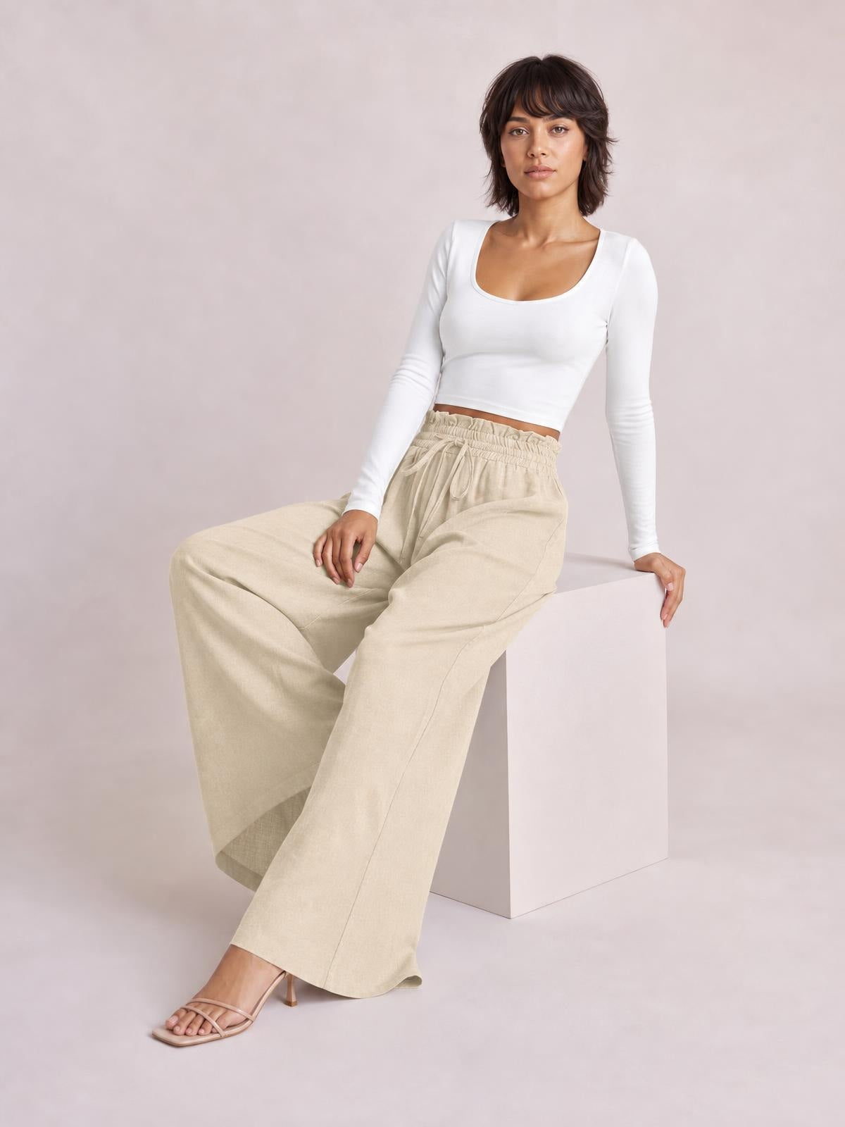 Model seated in beige drawstring wide leg pants with a relaxed, flowy silhouette, elasticized waist, visible pockets, and full-length hems.
