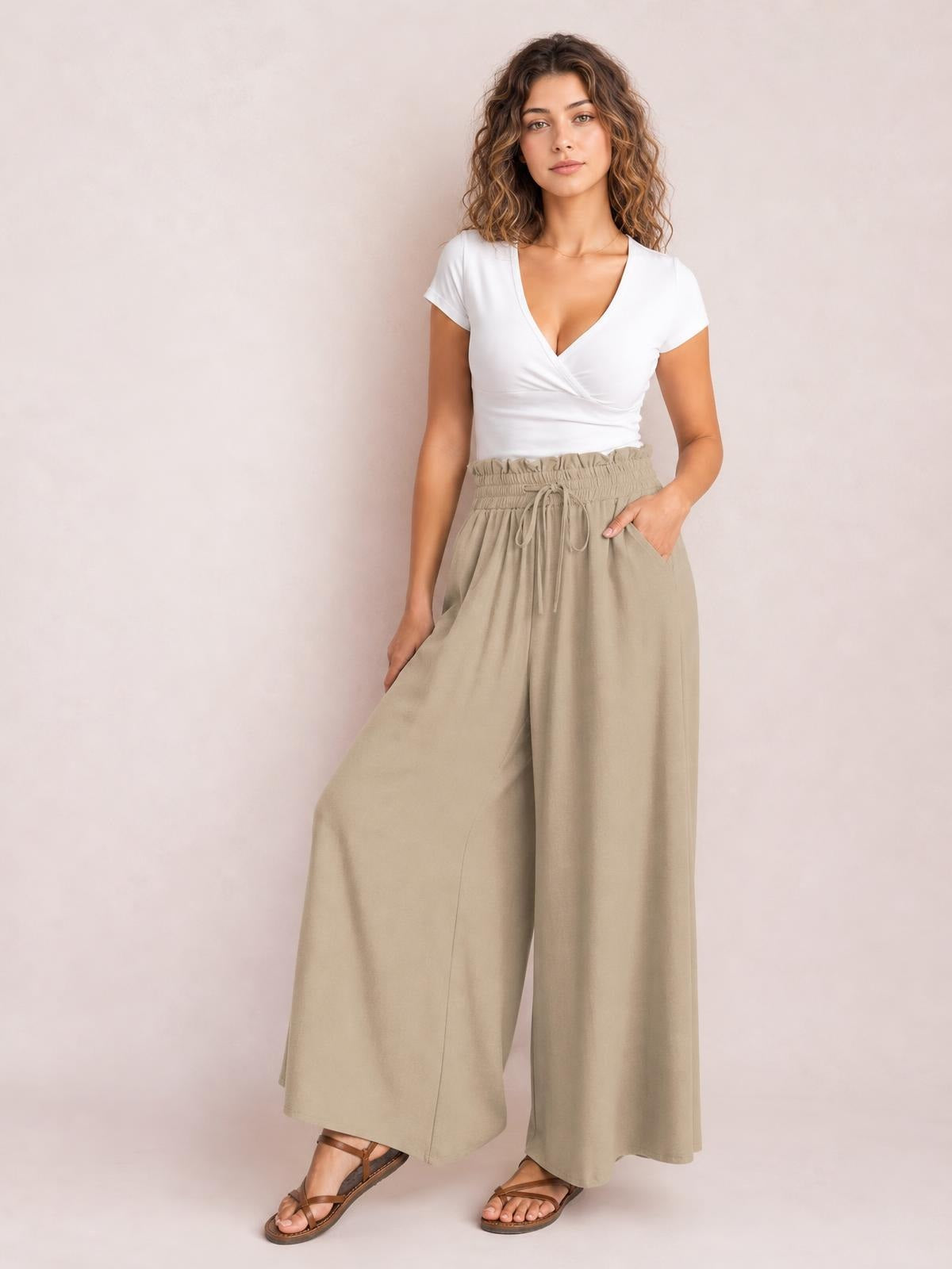 Model poses in beige drawstring wide-leg pants with a smocked waistband, side pockets, and a flowy full-length silhouette.