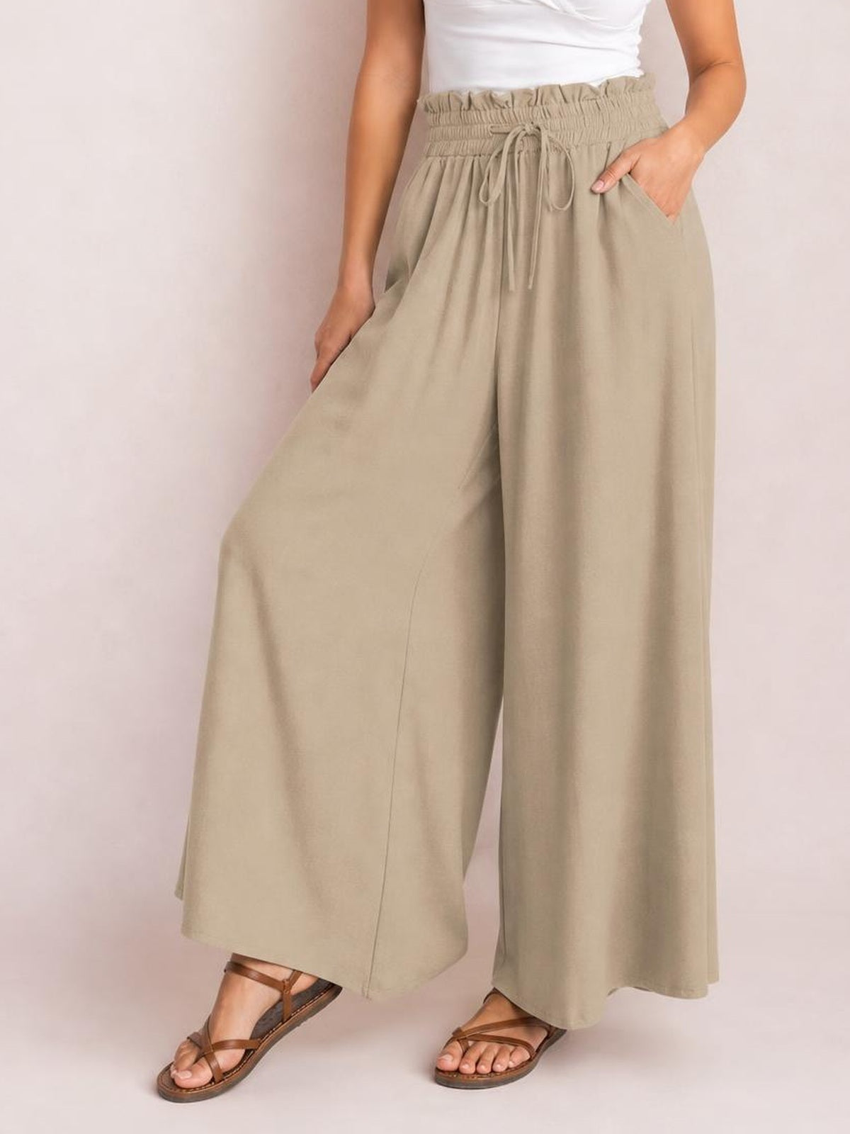 A model wears beige drawstring wide leg pants with a gathered waistband and side pockets, showing their relaxed, flowy silhouette while standing.