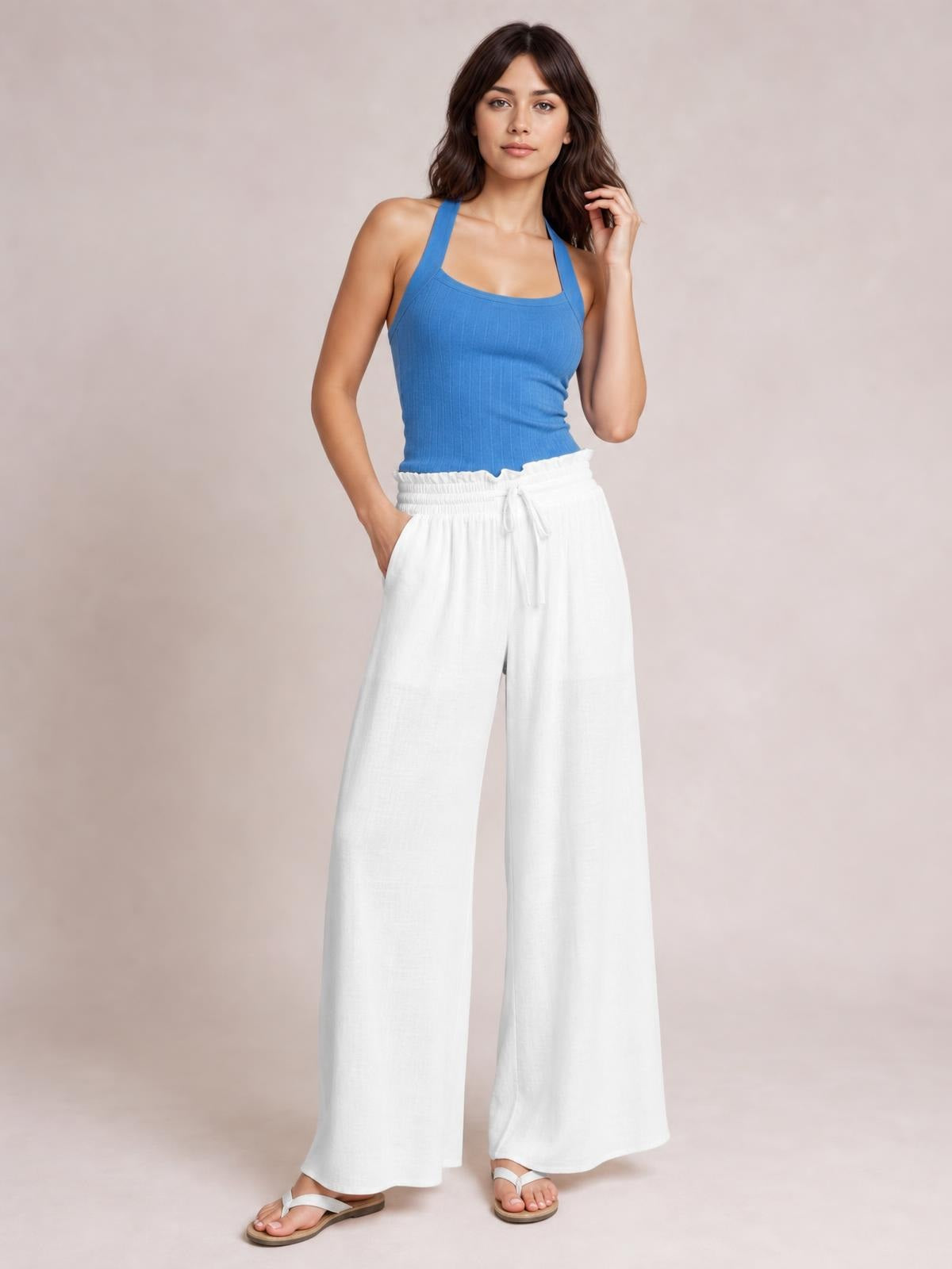 A model poses in white drawstring wide leg pants with a relaxed, flowy silhouette, gathered waistband, front tie, and side pockets, paired with sandals.