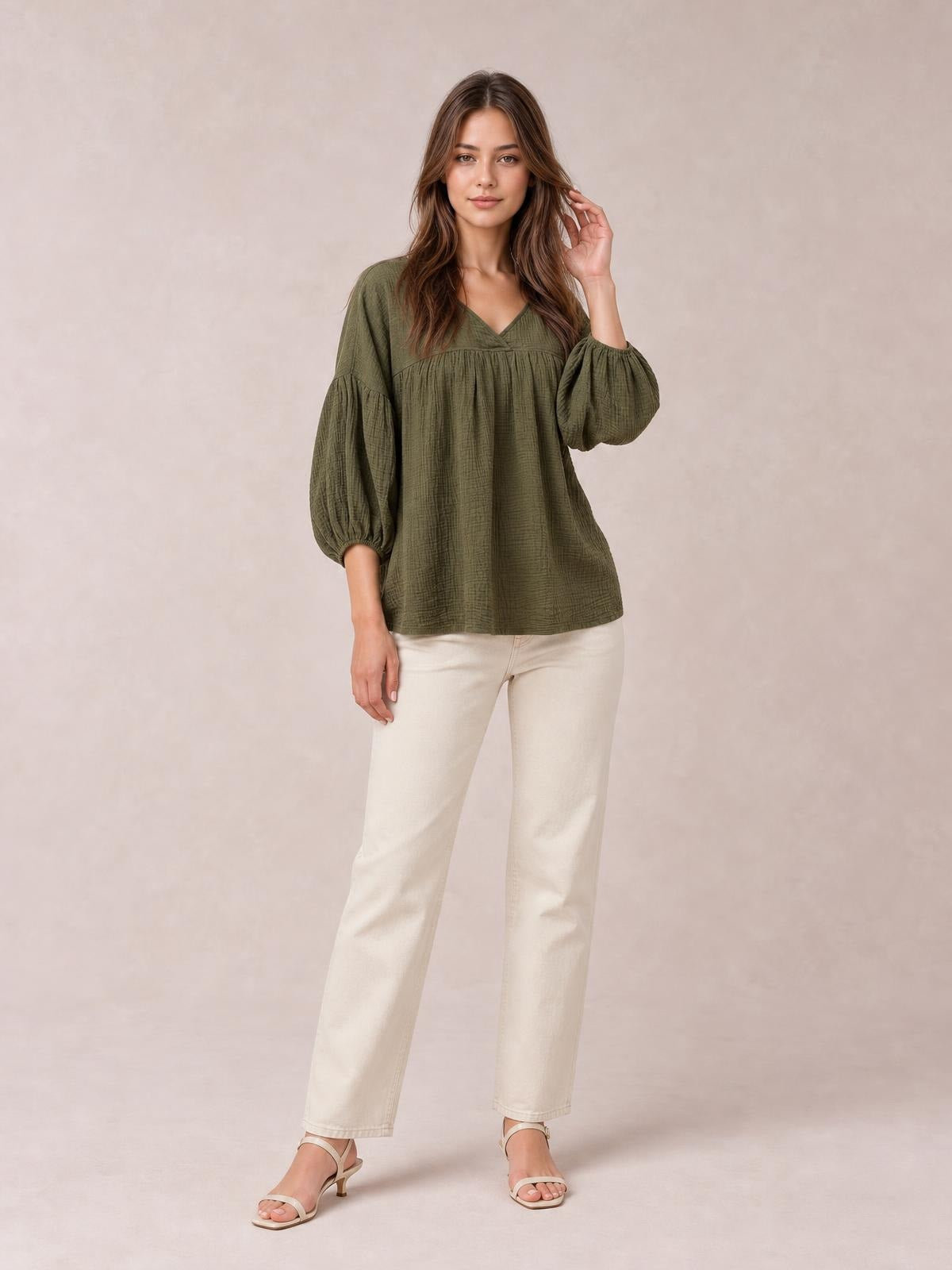 Model wears an olive green solid V-neck blouse with dropped shoulders, voluminous long sleeves, a gathered yoke, and a relaxed long silhouette while touching her hair.