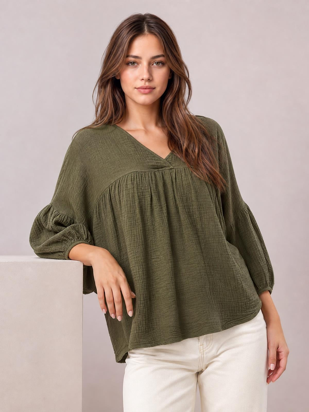A model leans on a block wearing an olive solid V-neck blouse with dropped shoulders, long gathered sleeves, and a relaxed, flowing silhouette.