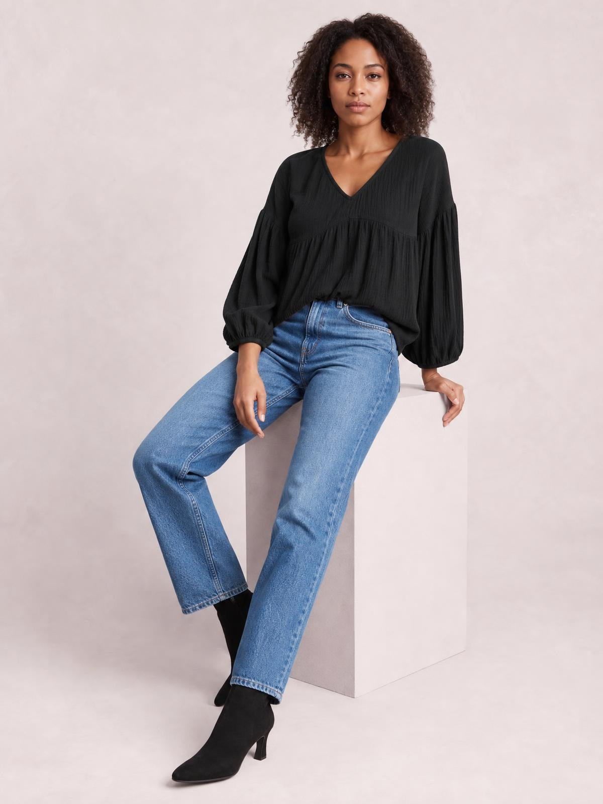 Model seated in a black solid blouse with a V-neck, dropped shoulders, long puff sleeves, gathered seams, and a loose long silhouette.