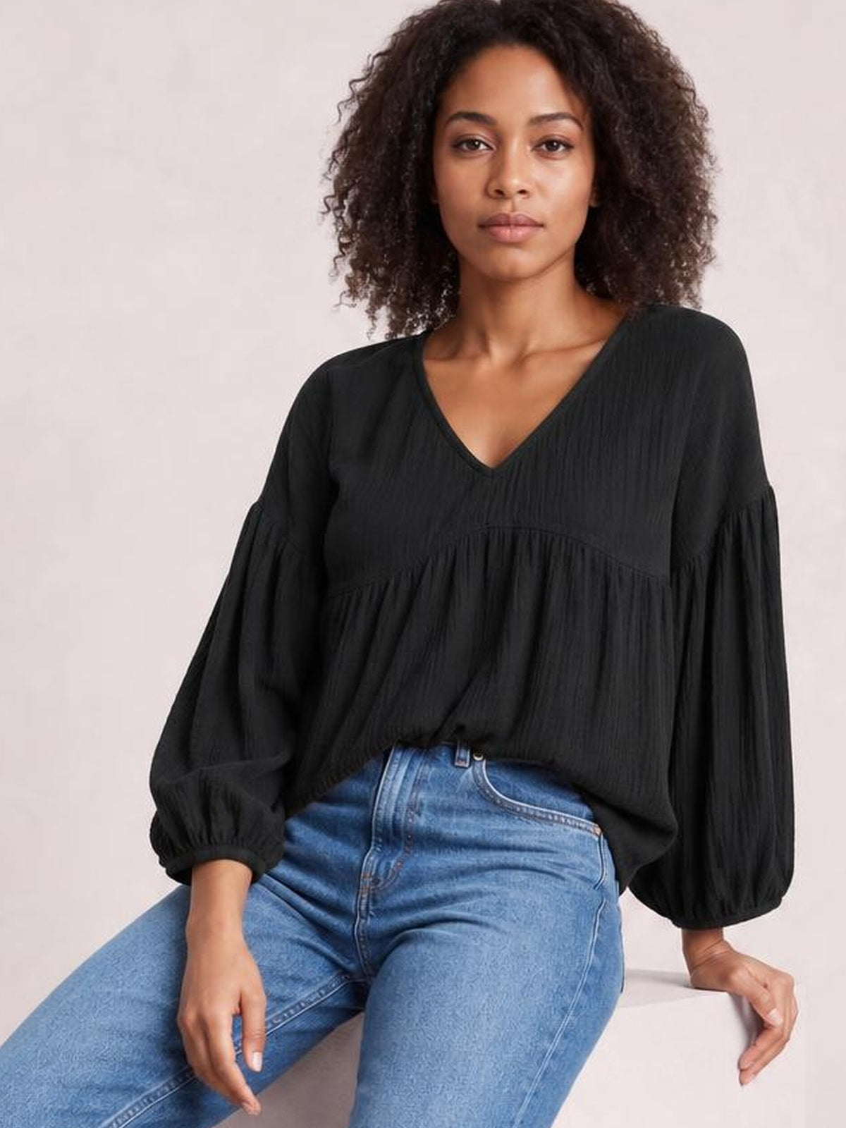 A model sits wearing a black dropped-shoulder V-neck blouse with long, gathered sleeves, elastic cuffs, a relaxed silhouette, and a tiered construction.