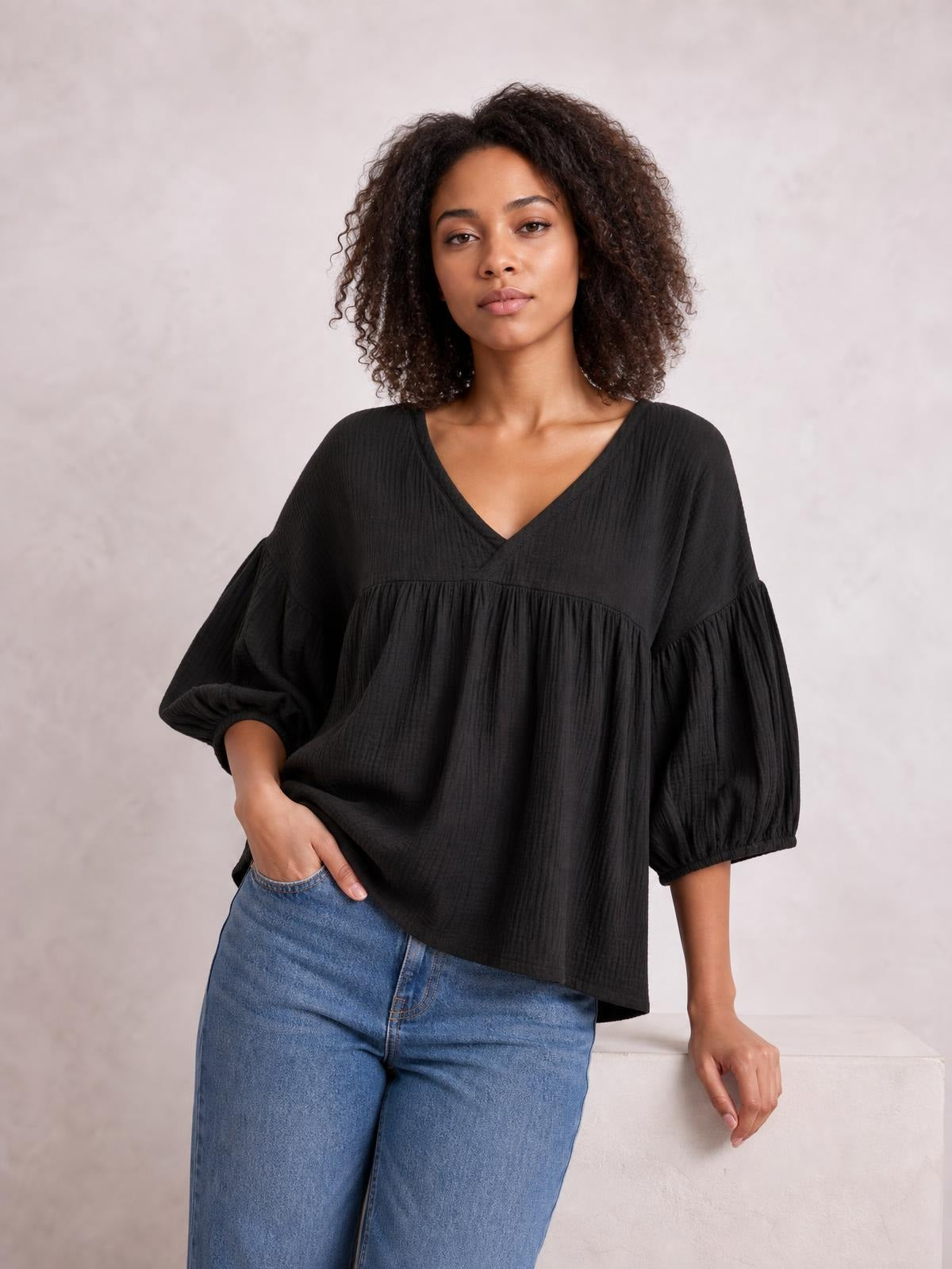 Model wears a solid black dropped shoulder V-neck blouse with a loose long silhouette, gathered three-quarter sleeves, and a tiered front.