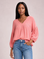 Model wearing a solid pink dropped shoulder V-neck blouse with a relaxed long silhouette, gathered seams, and voluminous long sleeves with elastic cuffs.
