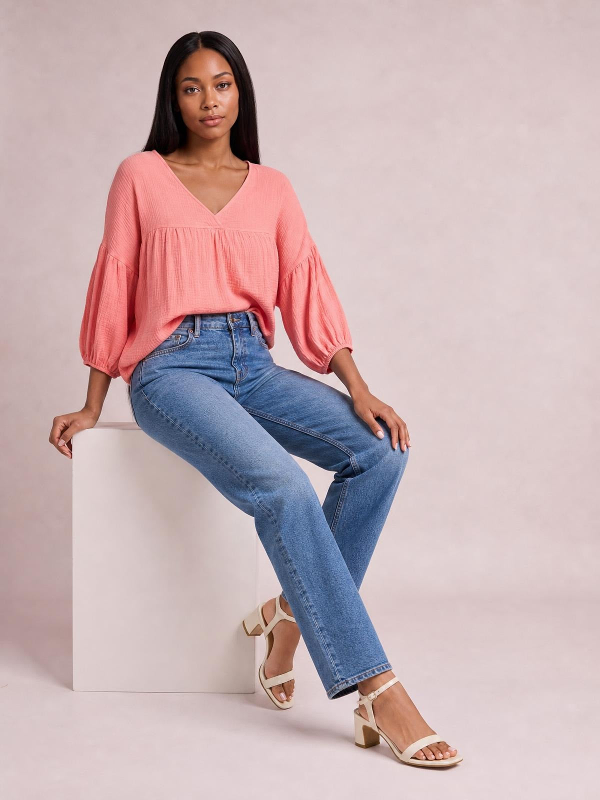 Model seated in a coral solid blouse with a V-neck, dropped shoulders, long billowy sleeves, gathered seams, and a relaxed long silhouette.