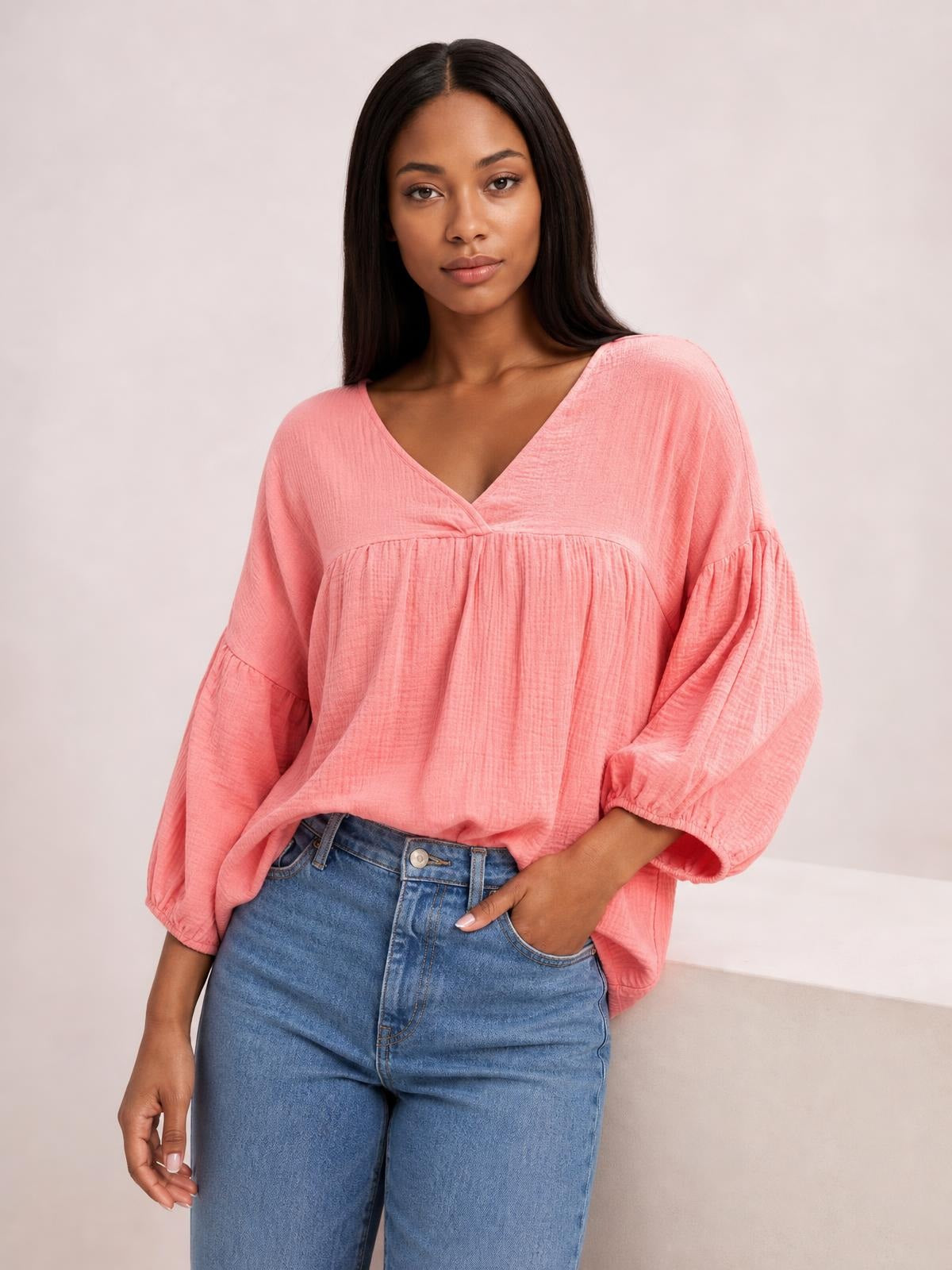 Model poses in a coral pink dropped-shoulder V-neck blouse with long balloon sleeves, gathered tiered construction, and a relaxed long silhouette.