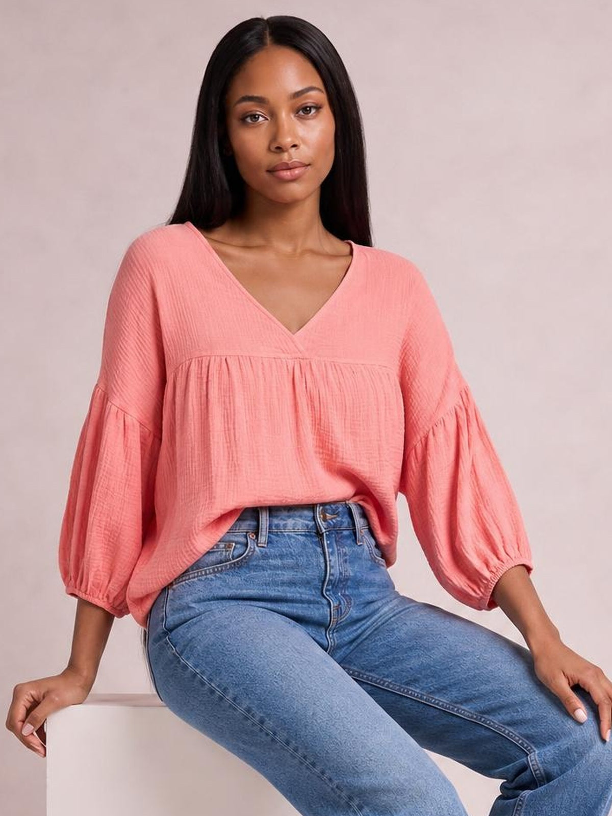 Model seated in a coral pink dropped shoulder V-neck blouse with a relaxed long silhouette, gathered tiered sleeves, and softly textured solid fabric.