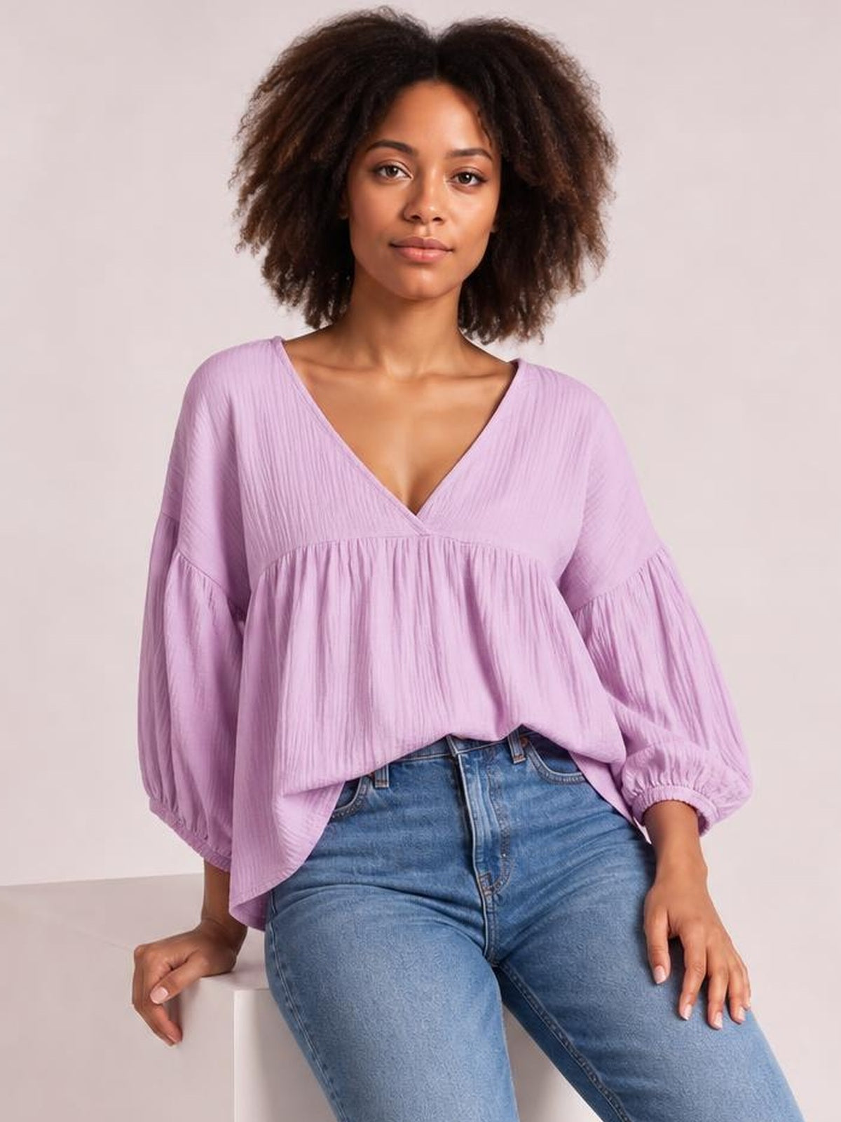 Model wearing a solid lavender dropped shoulder V-neck blouse with long balloon sleeves, gathered seams, and a relaxed flowing silhouette, seated on a stool.